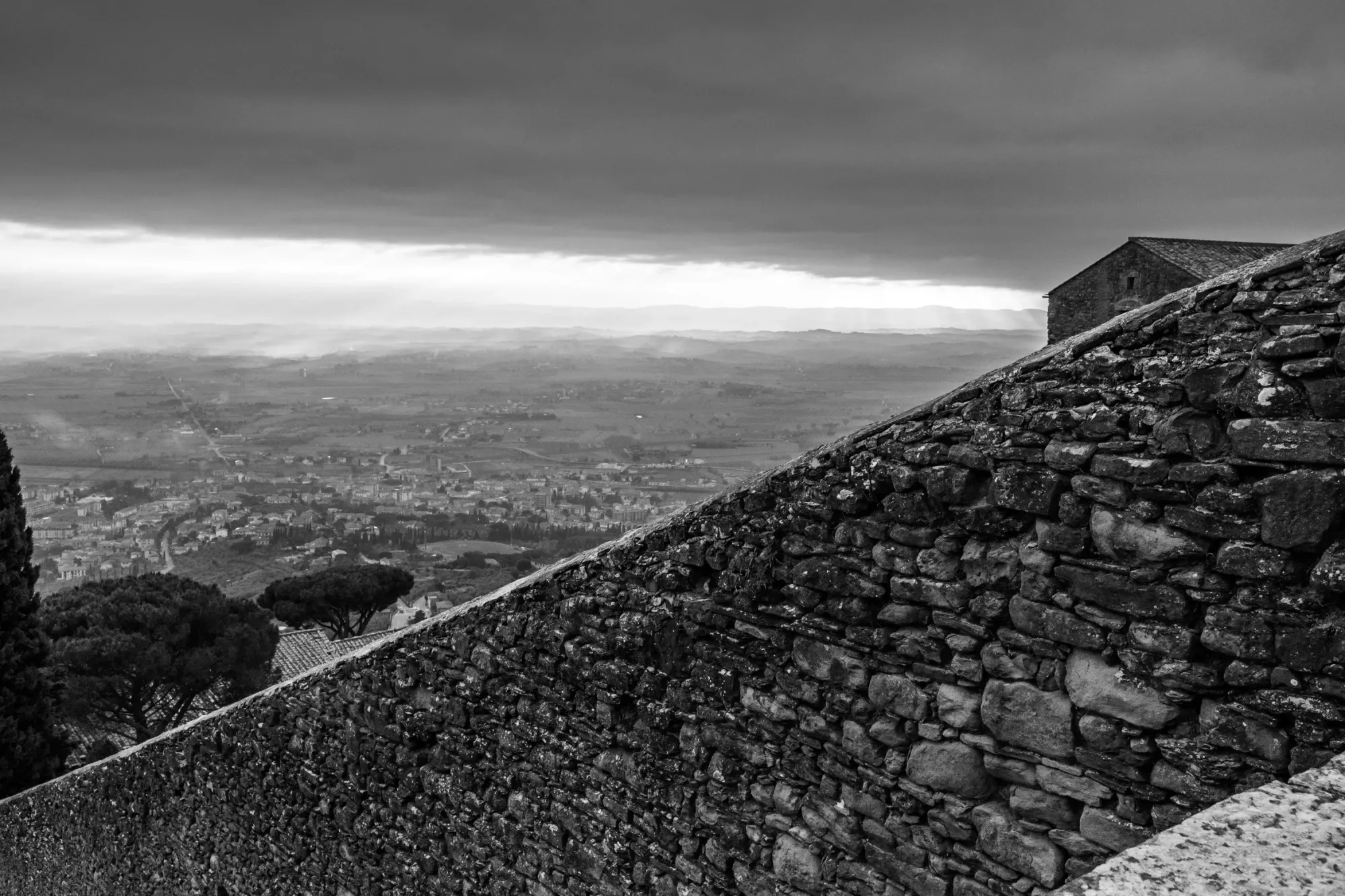 A monochromatic digital photograph of a large stone wall in Cortona, Italy. Beyond the wall is the Valdichiana valley and sunset breaking below low cloud cover. 