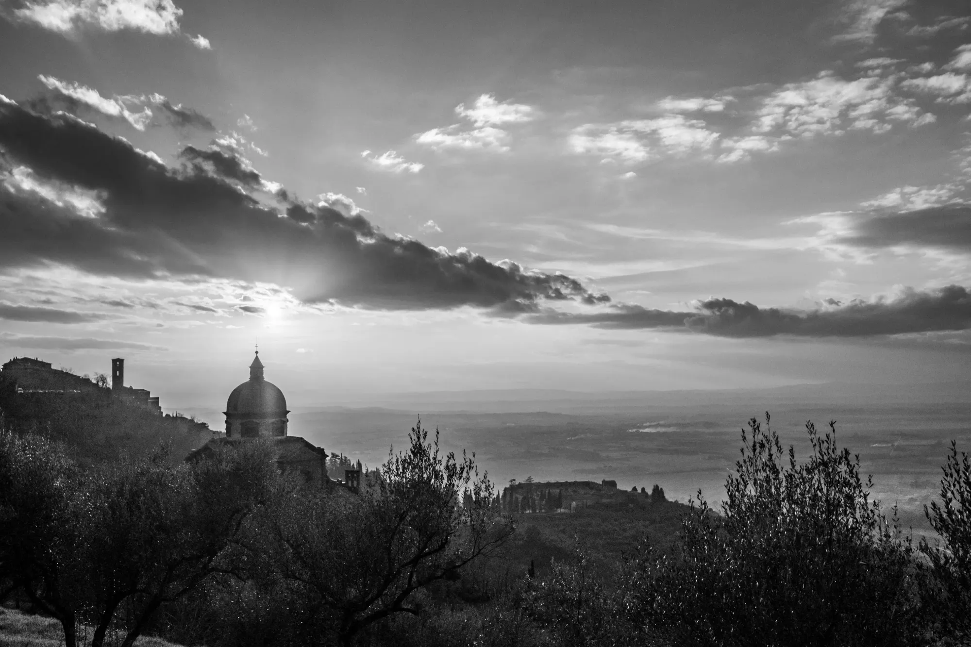 A monochromatic digital photograph of Chiesa di Santa Maria Nuova in Cortona Italy at sunset. The sky is filled with a backlit, abstract-shaped cloud.